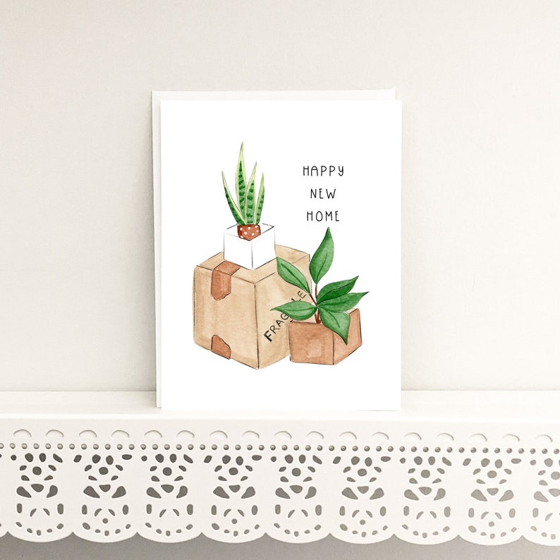 Peut inclure: Une carte blanche avec une illustration aquarelle de deux plantes en pot dans des bo&icirc;tes en carton. Le texte "Happy New Home" est &eacute;crit en lettres noires.