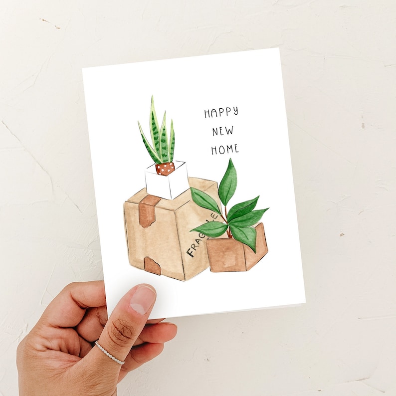 Peut inclure: Une carte de v&oelig;ux avec une illustration aquarelle de deux plantes en pot dans des bo&icirc;tes en carton. Le texte sur la carte est "Happy New Home".
