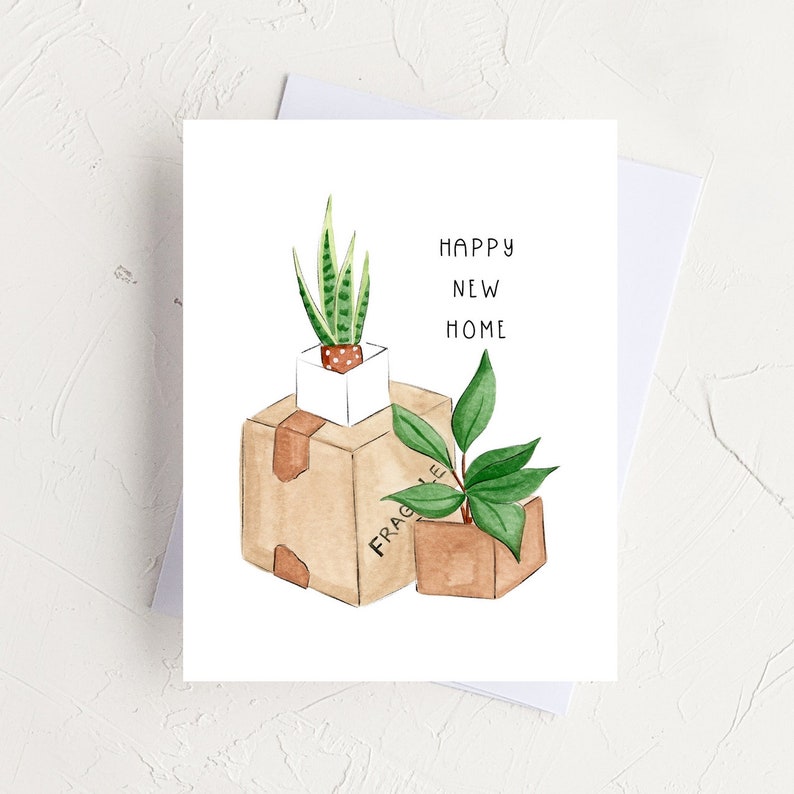 Peut inclure: Une carte de v&oelig;ux avec une illustration aquarelle de deux plantes en pot dans des bo&icirc;tes en carton. Le texte sur la carte est "Happy New Home".