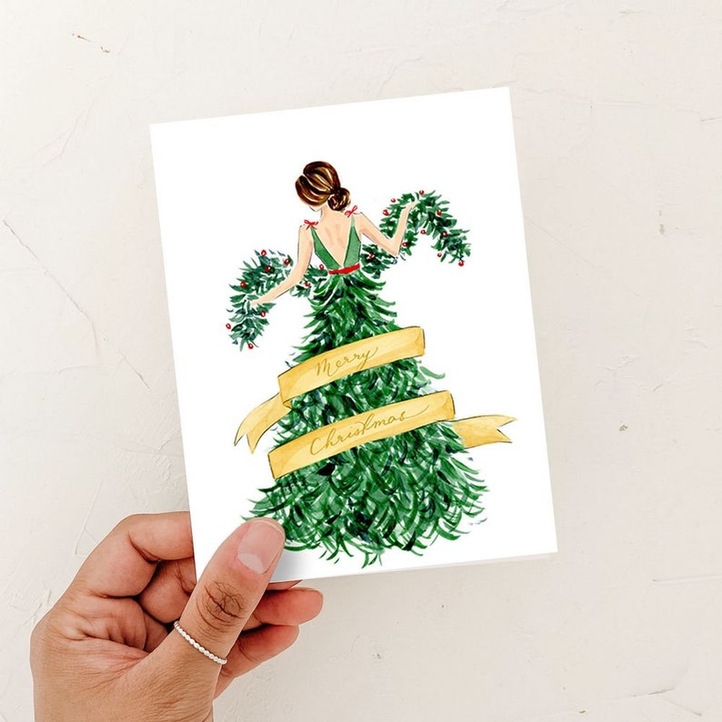 Op de afbeelding: Een aquarel-illustratie van een vrouw die een groene kerstboomjurk draagt met een gouden lint waarop "Merry Christmas" staat.