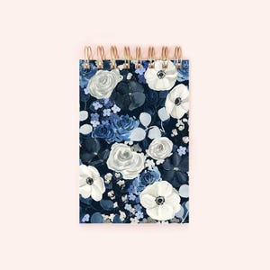 May include: A spiral-bound notebook with a floral design. The cover features white anemones, white and blue roses, and small blue flowers on a dark blue background. The spiral binding is gold-colored.