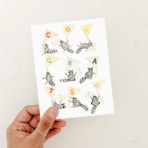 Peut inclure: Une carte de vœux blanche illustrée de plusieurs ratons laveurs dans diverses positions, chacun tenant un fanion triangulaire. Les drapeaux forment le mot CONGRATS avec des lettres colorées, accompagnés de petites illustrations de confettis.