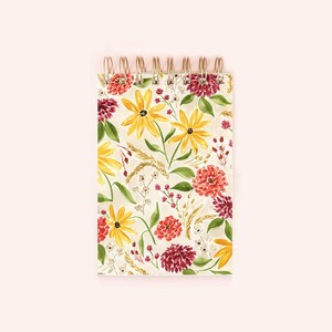 Peut inclure: Bloc-notes à spirale avec un motif floral. La couverture présente des tournesols jaunes, des fleurs rouges et oranges, des feuilles vertes et des épis de blé sur un fond crème. La reliure est dorée.