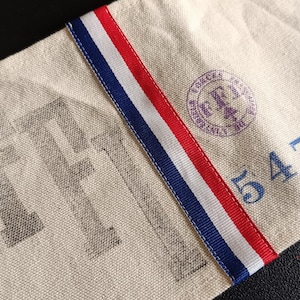 Puede incluir: Primer plano de una tela beige con las letras "FF" impresas en gris. Una cinta a rayas rojas, blancas y azules recorre la tela en diagonal. El número "5476" está impreso en azul, junto con un sello circular.