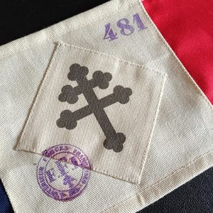 Può includere: Primo piano di una sezione di bandiera con una toppa beige con un disegno a croce scura. La bandiera ha sezioni rosse, bianche e blu. Il numero "481" è stampato in viola. È visibile anche un timbro viola.