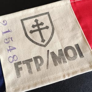 以下が含まれることがあります： 青、白、赤のパネルがある旗の一部をクローズアップ。白いパネルには「FTP/MOI」の文字と数字「21548」が表示されています。十字架の盾も表示されています。