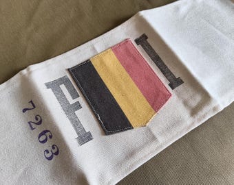 May include: A white canvas fabric strip featuring a central rectangular patch with vertical stripes in black, yellow, and red. The letter F is printed to the left of the patch, the letter I to the right, and the number 7263 is printed below the F.