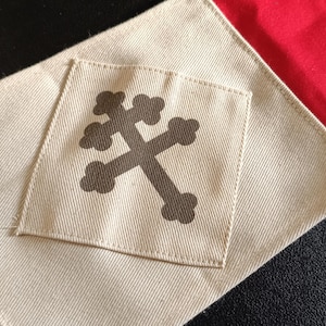 Peut inclure: Un patch en tissu avec une croix marron foncé sur un carré beige, cousu sur un tissu beige plus grand. Le patch est placé sur un fond de tissu rouge et bleu, créant une esthétique patriotique ou héraldique.