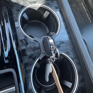 K&ouml;nnte beinhalten: Mittelkonsole eines Autos mit zwei Getr&auml;nkehaltern. Ein Schl&uuml;sselbund mit einem schwarzen Schl&uuml;sselanh&auml;nger und einem wei&szlig;en, handf&ouml;rmigen Anh&auml;nger liegt in einem Getr&auml;nkehalter. Ein Holzpaddel ist ebenfalls am Schl&uuml;sselbund befestigt.