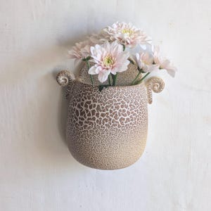 Pode incluir: Um vaso de parede com formato arredondado semelhante a um ovo e um acabamento texturizado e rachado em bege e branco. O recipiente possui duas pequenas alças decorativas curvas nas laterais e contém um buquê de flores de crisântemo rosa claro.