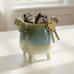 May include: A small, ceramic key holder with a blue and green glaze. The holder has three small legs and two curled handles. Several keys and key fobs are inside the holder, including car keys and house keys.