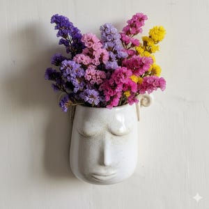 May include: A white ceramic wall vase shaped like a face, filled with colorful dried flowers. The flowers are a mix of purple, pink, and yellow. The vase has a matte finish and is mounted on a white wall.
