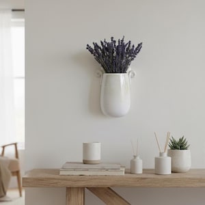 May include: A white ceramic wall vase with dried lavender, mounted on a white wall. A wooden table displays a candle, books, and small white ceramic vases with reeds and a potted succulent, creating a minimalist aesthetic.