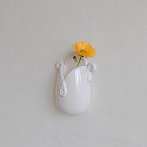May include: A white ceramic wall vase with a single yellow flower. The vase has a unique, curved design with decorative swirls and a glossy finish. The flower's stem is visible inside the vase.