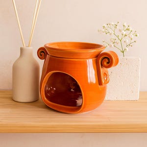 May include: An orange ceramic oil warmer with a large opening and decorative scroll handles. A beige diffuser bottle with reeds and a small white floral arrangement are also present on a wooden shelf.