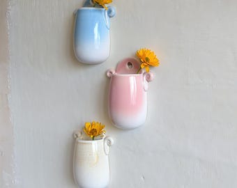 May include: Three ceramic wall vases in blue, pink, and cream, each with a white ombre effect at the base. The vases have a small hole for hanging and are adorned with yellow flowers. The vases are a decorative home decor item.