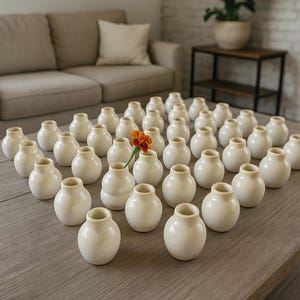 May include: A display of small, cream-coloured ceramic vases on a wooden table. One vase contains an orange flower. The vases are round with a narrow neck. The background features a beige sofa and a side table.