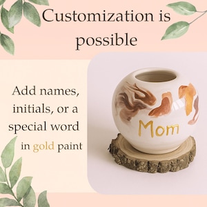 May include: A white ceramic vase with brown and gold abstract designs and the word "Mom" in gold paint. The vase sits on a small wooden slice. Text on the left says "Customisation is possible" and "Add names, initials, or a special word in gold paint."