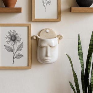 Può includere: Un vaso da parete in ceramica bianca a forma di viso con gli occhi chiusi. Il vaso ha una parte superiore beige e riccioli decorativi su ogni lato. È montato su una parete bianca, vicino a opere d'arte botaniche incorniciate e una pianta in vaso.