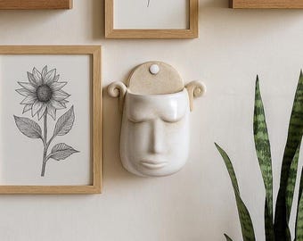 以下が含まれることがあります： 目を閉じた顔の形をした白いセラミック製の壁掛けプランター。 プランターはベージュのトップと両側に装飾的なカールが付いています。 白い壁に取り付けられており、額入りの植物画と鉢植えの植物の近くにあります。