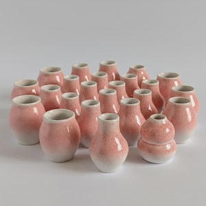 May include: A collection of small, pink ceramic vases with a speckled texture. The vases have a variety of shapes, including round, cylindrical, and gourd-like forms. The bases of the vases are white, creating a gradient effect.