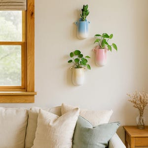Op de afbeelding: Drie aan de muur gemonteerde plantenbakken in een kleurengradiënt: blauw, roze en geel. Elke plantenbak bevat een andere groene plant. De plantenbakken hangen aan een neutraal gekleurde muur boven een crèmekleurige bank met kussens en een houten bijzettafel.