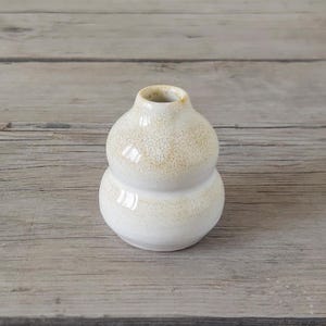 Può includere: Un vaso in ceramica bianca con una glassa macchiettata. Il vaso ha una forma arrotondata, simile a una zucca, con due rigonfiamenti distinti.