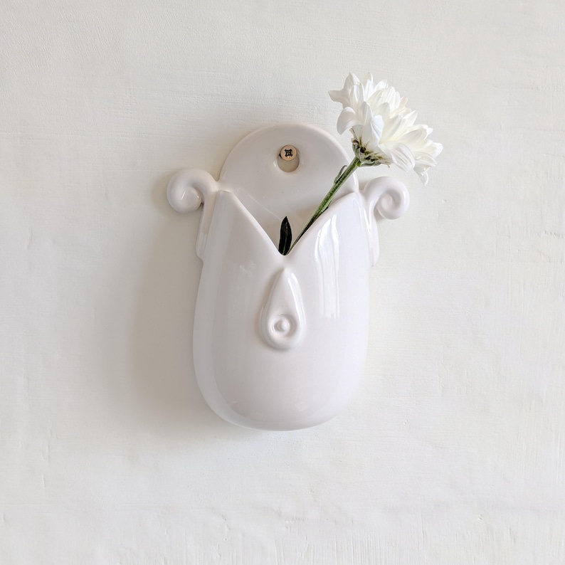 May include: A white ceramic wall vase with a distinctive design, featuring a central opening and decorative swirls. A white flower with green stem is placed inside the vase. The vase is mounted on a white wall.