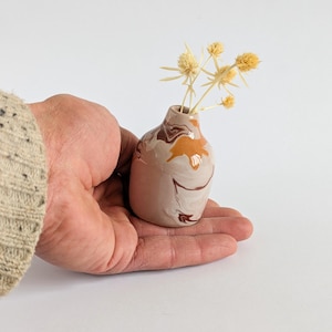 Könnte beinhalten: Eine kleine Keramikvase mit Marmormuster in Braun-, Beige- und Orangetönen, die getrocknete gelbe Blumen enthält. Die Vase wird in einer Hand vor weißem Hintergrund gehalten. Die Vase ist etwa 7,6 cm hoch.