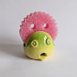 Op de afbeelding: Een grillige keramische spons houder in de vorm van een verbaasd gezicht, met een roze spons erin. De houder is voornamelijk geel en groen, met gestileerde oog- en monddetails. Een leuk en functioneel keukenaccessoire.