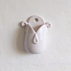 May include: A white ceramic wall pocket, shaped like a tulip. It features a rounded top with a hanging hole, a central opening, and curled decorative elements. The surface is smooth and glossy, with a clean, minimalist design.