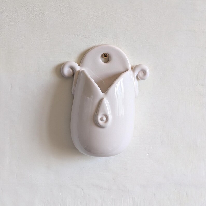May include: A white ceramic wall pocket, shaped like a tulip. It features a rounded top with a hanging hole, a central opening, and curled decorative elements. The surface is smooth and glossy, with a clean, minimalist design.