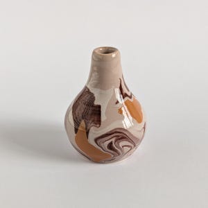 Könnte beinhalten: Kleine, dekorative Vase mit bauchigem Körper und schmalem Hals. Die Vase hat ein marmoriertes Design in Braun-, Beige- und Orangetönen. Die Vase ist etwa 13 cm hoch und eignet sich zur Präsentation kleiner Blumen oder als dekoratives Element.