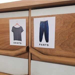 Peut inclure: Deux étiquettes blanches avec du texte noir sur une commode en bois. L'étiquette de gauche indique "CHEMISES" et montre un t-shirt gris sur un cintre. L'étiquette de droite indique "PANTALONS" et montre un pantalon en jean bleu sur un cintre.