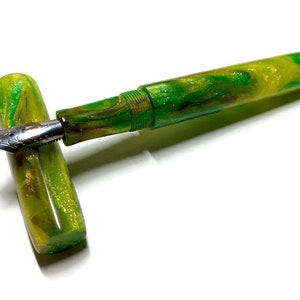 Puede incluir: Una pluma estilogr&aacute;fica verde y dorada con acabado brillante. La pluma est&aacute; cerrada y la punta es visible.