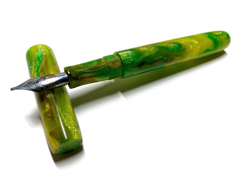 Puede incluir: Una pluma estilogr&aacute;fica verde y dorada con acabado brillante. La pluma est&aacute; cerrada y la punta es visible.