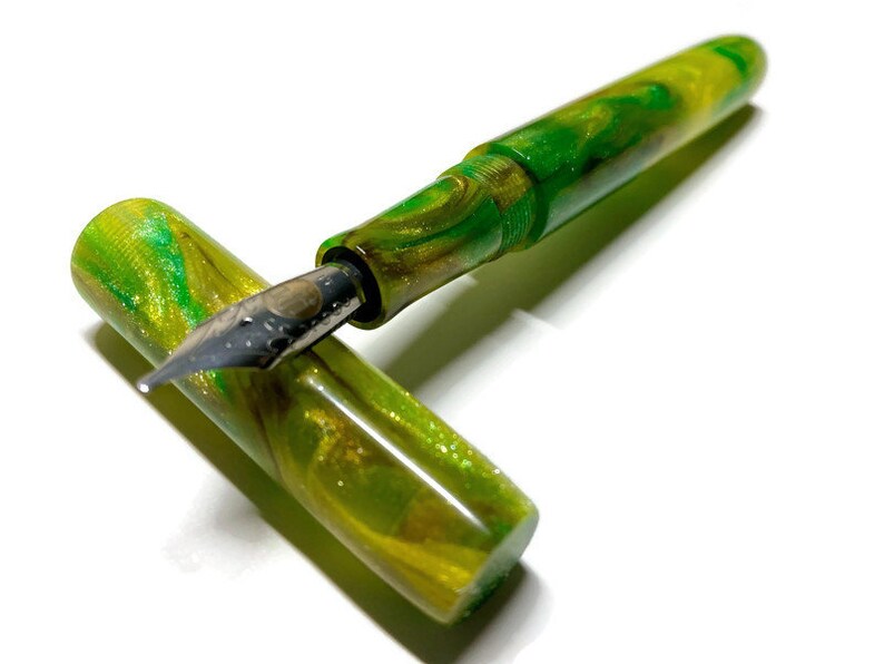 Puede incluir: Una pluma estilogr&aacute;fica verde y dorada con una plumilla de metal. La pluma est&aacute; hecha de resina marmoleada con un acabado brillante.