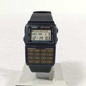 May include: Black Casio Data Bank watch with a calculator keypad. The digital display shows the time and date. The watch is on a white display stand with the Casio logo.