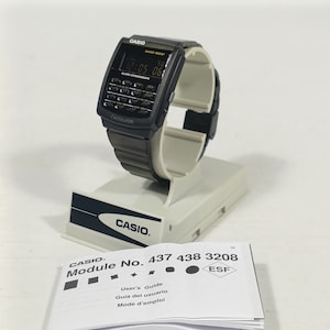May include: A black Casio digital watch with calculator functions, displayed on a white stand. The watch face shows the time and day. The watch has a black band. Included are the user guide and a Casio tag.