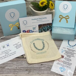 May include: A light blue box with a white and gold bow on top. The box is labeled "DawnsBeads Designs". The box is sitting on a wooden surface. There is a small white card with the text "Spend $20.00 Use Coupon Code SAVES Save $5.00 your next visit! *no valid on Sale items*" and a small white card with the text "Thank you! for Supporting My Small Business!"  There is also a small white card with the text "Handmade Bead Jewelry And Accessories www.etsy.com/shop/dawnsbeaddesigns" and a small white card with the text "Jewelry Care Remove jewelry before bed Remove jewelry before applying perfume, cologne or lotions Remove jewelry before bathing or exercise Store jewelry flat in box, bag or drawer Clean Jewelry with any basic costume jewelry cleaner With Proper Care, storage and wear your jewelry will continue to shine".