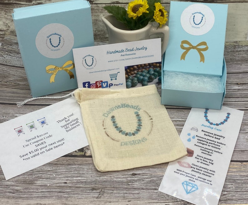 May include: A light blue box with a white and gold bow on top. The box is labeled "DawnsBeads Designs". The box is sitting on a wooden surface. There is a small white card with the text "Spend $20.00 Use Coupon Code SAVES Save $5.00 your next visit! *no valid on Sale items*" and a small white card with the text "Thank you! for Supporting My Small Business!"  There is also a small white card with the text "Handmade Bead Jewelry And Accessories www.etsy.com/shop/dawnsbeaddesigns" and a small white card with the text "Jewelry Care Remove jewelry before bed Remove jewelry before applying perfume, cologne or lotions Remove jewelry before bathing or exercise Store jewelry flat in box, bag or drawer Clean Jewelry with any basic costume jewelry cleaner With Proper Care, storage and wear your jewelry will continue to shine".