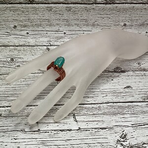 May include: A copper wire ring with a teal green stone set in the center. The ring is on a white mannequin hand.