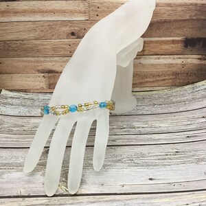 Puede incluir: Un brazalete de cuentas azules y doradas. El brazalete está en un maniquí de mano blanco.