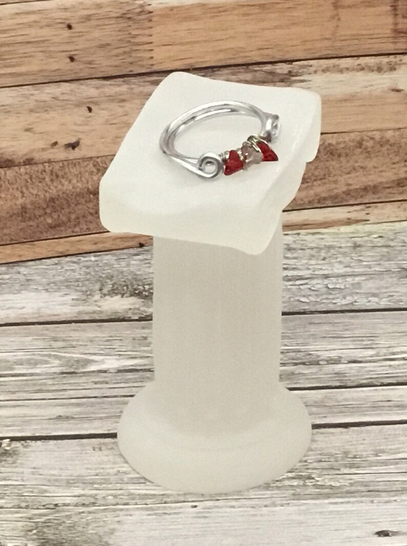 May include: A silver wire ring with three red and silver beads. The ring is displayed on a white pedestal.