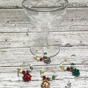 Peut inclure: Trois breloques en verre à bordure dorée avec des fils de perles colorés et des décorations de nœuds en rouge, vert et or. Les breloques sont conçues pour être accrochées aux verres à vin.