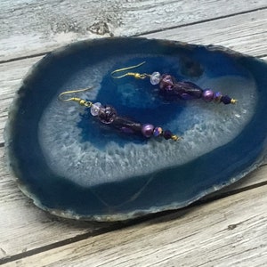 Pode incluir: Um par de brincos pendentes roxos e dourados com contas de vidro e um detalhe de pérola. Os brincos são exibidos em uma fatia de ágata azul.