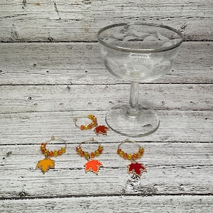 May include: Four glass charms with gold rings and colorful fall leaf designs. The charms are on a wooden surface with a clear glass goblet in the background.