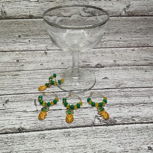 Peut inclure: Quatre charmes en verre en forme d'ananas dorés avec des perles vertes et jaunes. Les charmes sont suspendus à un gobelet en verre.