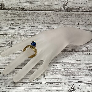 May include: A gold ring with a blue glass stone set in the center. The ring is on a white mannequin hand.