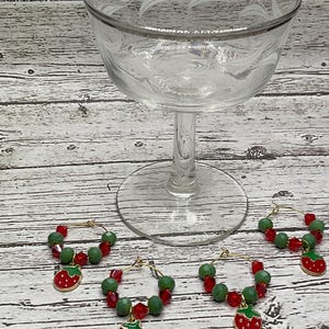 Peut inclure: Quatre charmes de verre à vin dorés avec des perles de verre rouges et vertes et un charme de fraise rouge. Les charmes sont sur une surface en bois blanc.